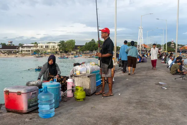 Bali, Endonezya - 22 Aralık 2024: Bir kadın önünde otururken erkek bir balıkçı oltasıyla iskelede duruyor. Bir grup insan yanımızdan geçiyor. Sahne canlı ve hareketli.