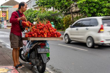 Bali, Endonezya - 10 Aralık 2024: Adam motosiklet üzerinde rambutan adı verilen meyveler satıyor. Meyve kırmızıdır ve onlardan çok var.