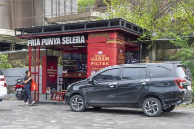 Bali, Endonezya - 10 Aralık 2024: Üzerinde PRIA PUNYA SELERA, Gudang Garam Filtresi yazan kırmızı bir binanın önünde yürüyen kadın