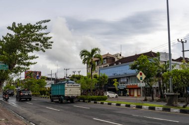Bali, Endonezya - 10 Aralık 2024: Yoğun bir cadde ve bir kamyon geçiyor. Sokak ağaçlar ve binalarla dolu. Gökyüzü bulutlu ve güneş görünmüyor.