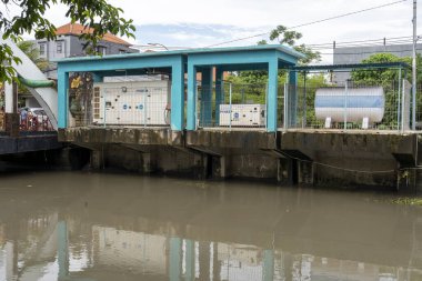 Bali, Endonezya - 28 Aralık 2024: Arkasında yeşil bir bina olan bir nehir üzerinde köprü. Köprü yapraklarla kaplı ve su bulanık.