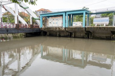 Bali, Endonezya - 28 Aralık 2024: Arkasında yeşil bir bina olan bir nehir üzerinde köprü. Köprü yapraklarla kaplı ve su bulanık.