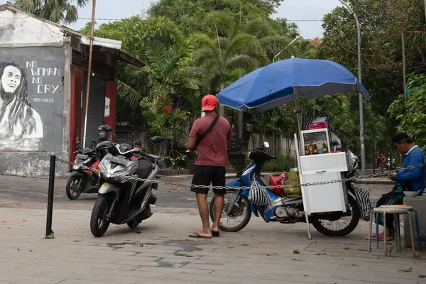 Bali, Endonezya - 11 Aralık 2024: Adam yemek arabasının yanında mavi bir şemsiye altında duruyor. Yakınlarda iki motosiklet var.