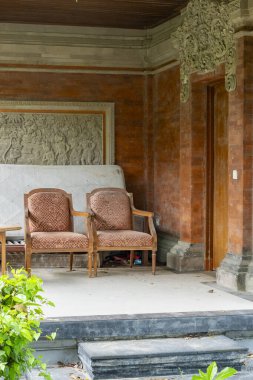 Bali, Endonezya - 17 Aralık 2024: Tahta veranda, iki sandalye ve bir bank. Sandalyeler kahverengi ve bank beyaz. Veranda ağaçlar ve çalılarla çevrili.