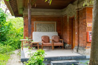Bali, Endonezya - 17 Aralık 2024: Tahta veranda, iki sandalye ve bir bank. Sandalyeler kahverengi ve bank beyaz. Veranda ağaçlar ve çalılarla çevrili.