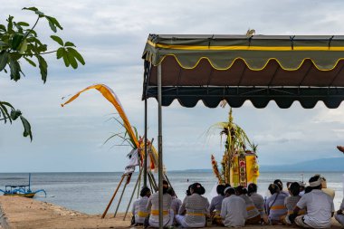 Bali, Endonezya - 17 Aralık 2024: Bir grup insan sahilde bir gölgelik altında oturup dua ediyor. İnsanlar beyaz giyinmiş ve kumların üzerinde oturuyorlar. Manzara huzurlu ve rahatlatıcı.