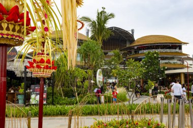 Bali, Endonezya - 17 Aralık 2024: Icon Mall Bali. Bir grup insan arka planda bir binayla parkta yürüyorlar. Manzara sakin ve huzurlu.