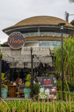 Bali, Endonezya - 17 Aralık 2024: Icon Mall Bali. Bir grup insan arka planda bir binayla parkta yürüyorlar. Manzara sakin ve huzurlu.