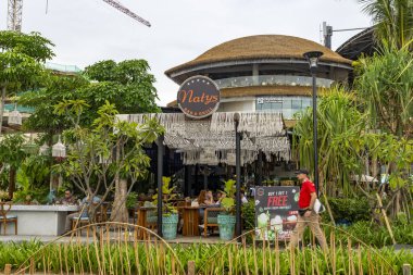 Bali, Endonezya - 17 Aralık 2024: Icon Mall Bali. Bir grup insan arka planda bir binayla parkta yürüyorlar. Manzara sakin ve huzurlu.