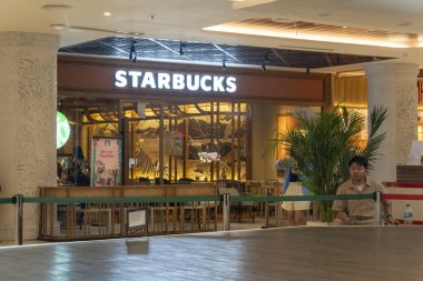 Bali, Endonezya - 17 Aralık 2024: Bali 'deki Icon Bali Alışveriş Merkezi. Starbucks mağazasının önünde bir adam oturuyor. Burası açık ve kapısında yeşil bir tabela var.