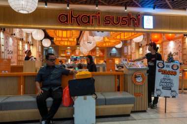 Bali, Endonezya - 17 Aralık 2024: Bali 'deki Icon Bali Alışveriş Merkezi. Adam Akari suşi restoranının önündeki bankta oturuyor. Arkasında bir kadın duruyor, ona menüyü uzatıyor.