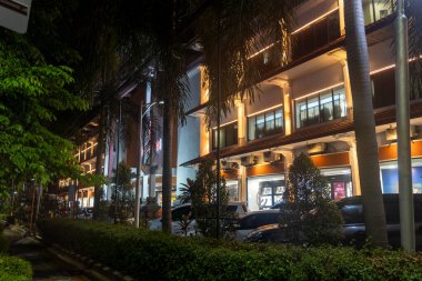 Bali, Endonezya - 17 Aralık 2024: Ağaçlı ve palmiye ağaçlı bir alışveriş merkezinin gece manzarası. Alışveriş merkezi aydınlandı ve park yerinde bir sürü araba var.