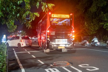 Bali, Endonezya - 17 Aralık 2024: Otobüs gece bir caddeden geçiyor. Otobüs kırmızı ve beyazdır. Sokaklar arabalar ve motosikletlerle dolu.