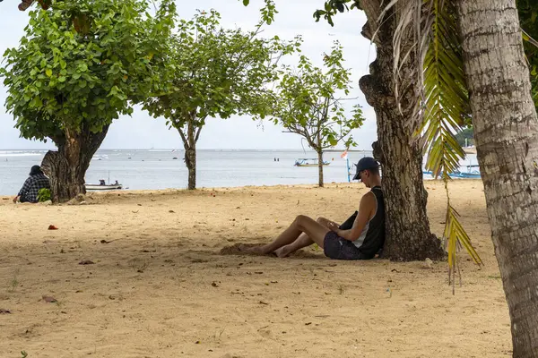 Bali, Endonezya - 17 Aralık 2024: İnsanlar sahilde bir ağacın altında oturuyor. Kumsal boş ve adam telefon okuyor.