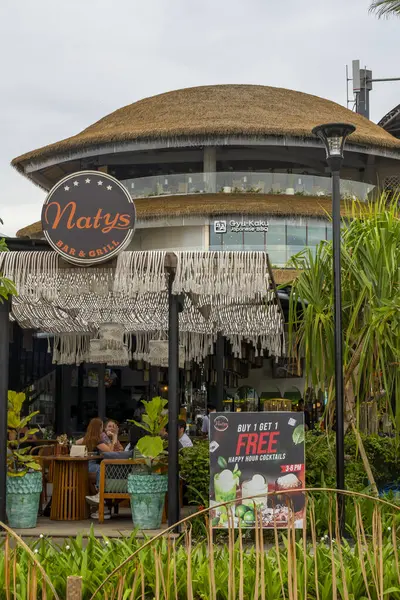 Bali, Endonezya - 17 Aralık 2024: Icon Mall Bali. Bir grup insan arka planda bir binayla parkta yürüyorlar. Manzara sakin ve huzurlu.