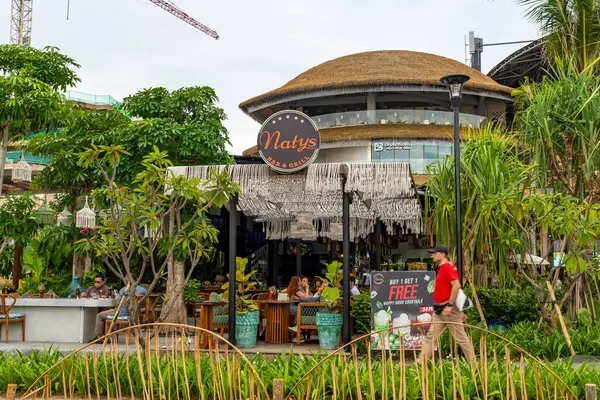 Bali, Endonezya - 17 Aralık 2024: Icon Mall Bali. Bir grup insan arka planda bir binayla parkta yürüyorlar. Manzara sakin ve huzurlu.