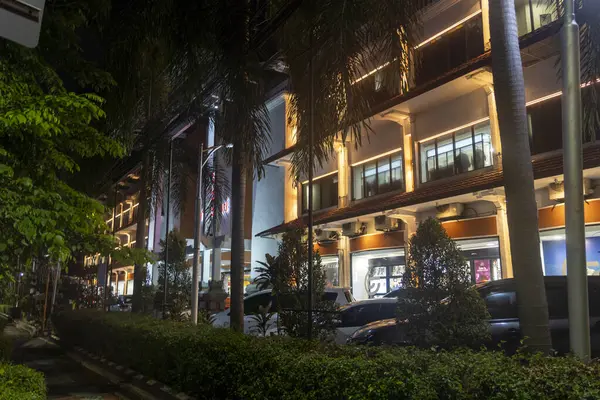 Bali, Endonezya - 17 Aralık 2024: Ağaçlı ve palmiye ağaçlı bir alışveriş merkezinin gece manzarası. Alışveriş merkezi aydınlandı ve park yerinde bir sürü araba var.