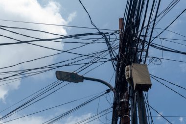 Bulutlu bir gökyüzüne karşı yıpranmış elektrik ve iletişim kablolarından oluşan kaotik bir ağ, kentsel altyapı sorunlarını vurguluyor..