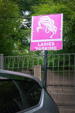 Üzerinde kadınlar için park yeri yazan pembe bir tabela. İşaret, otoparktaki çitin yanındaki direkte.