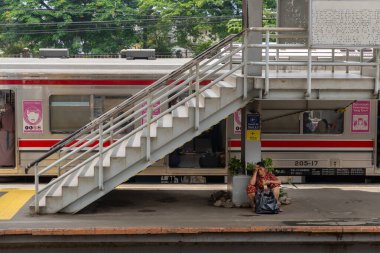 Jakarta, Endonezya - 9 Şubat 2025: Kadın siyah plastik bir çantayla tren peronunda oturuyor. Tren istasyonda park halinde ve pembe beyaz bir tasarımı var.