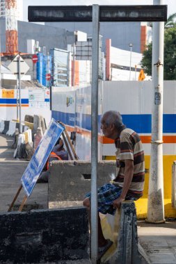 Jakarta, Endonezya - 9 Şubat 2025: Adam bir tabelanın yanındaki çimento bloğunda oturuyor. Adam çizgili gömlek ve mavi şort giyiyor.