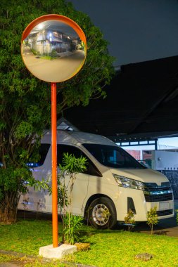 Beyaz minibüs aynanın önüne park edilmiş. Ayna turuncu ve minibüsü yansıtıyor. Sahne bir otoparkta geçiyor.