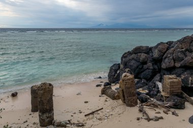 Arka planda büyük bir su kütlesi olan Rocky plajı. Kayalar sahilin etrafına saçılmış ve kumların üzerinde tahta bloklar var. Huzurlu bir sahne.