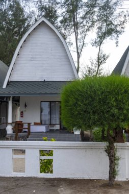 Beyaz dış görünüşlü ve yeşil çatılı bir ev. Evin önünde yeşil çalılar olan beyaz bir çit var.