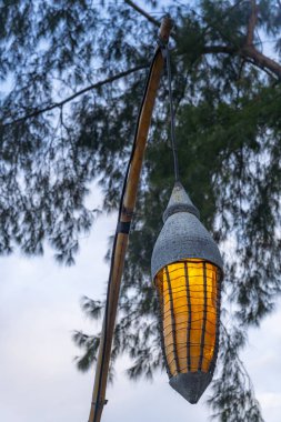 Arka planda mavi bir şemsiyeyle bambu direğine asılı bir lamba. Lamba yanıyor ve şemsiye açık.