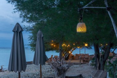 Şemsiyeli plaj sahnesi ve ağaçtan sarkan lambalı bir masa. Manzara sakin ve huzurlu.