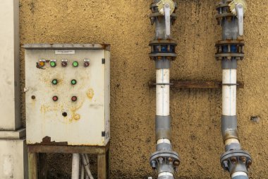 Bali, Endonezya - 23 Aralık 2024: Üzerinde yeşil ışık olan paslı kutu iki borunun yanında duruyor. Kutu eski ve yıpranmış, borular da paslı ve kirli. Sahne ihmalkarlık ve çürümedir.