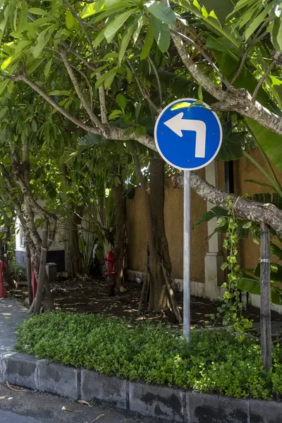 Solu gösteren beyaz oklu mavi işaret. Tabela ağaçlar ve çalılarla dolu bir parkta.