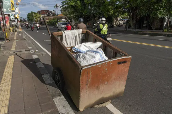 Adam üzerinde torba olan bir arabayı itiyor. Araba caddede ve etrafında başka insanlar ve araçlar var.