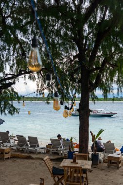 Gili, Lombok, Endonezya - 3 Ocak 2025: Suda bir ağaç ve bir tekne olan plaj sahnesi. İnsanların oturup dinlenmesi için kurulmuş birkaç sandalye ve masa var. Manzara huzurlu ve rahatlatıcı.