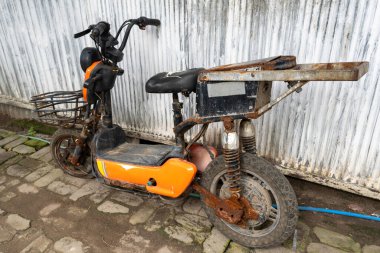 Önünde sepeti olan paslı eski bir scooter. Sepet boş. Scooter duvara yaslanmış.