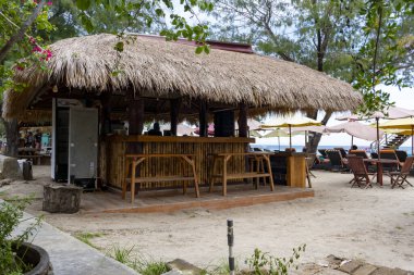 Gili, Lombok, Endonezya - 3 Ocak 2025: Sazdan çatılı küçük bir kulübe sahilde bulunuyor. Kulübe bir taş duvar ve birkaç bankla çevrili. Huzur dolu bir sahne.