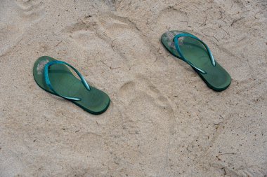 İki tane parmak arası terlik kumun üzerinde yatıyor. Kum beyaz ve parmak arası terlikler yeşil.
