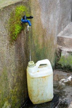 Beyaz plastik kap bir musluğun yanında. Konteynır suyla dolu ve kovayı doldurmak için kullanılıyor.