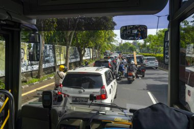 Bali, Endonezya - 19 Aralık 2024: Otobüs kalabalık bir caddede birçok araba ve motosikletle ilerliyor. Otobüs şoförü pencereden dışarı bakıyor ve trafiği izliyor. Sahne kalabalık ve hareketli.