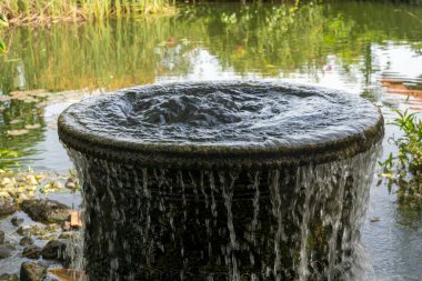 Büyük, yuvarlak, içinden su akan siyah bir kap. Su bir gölete dökülüyor.