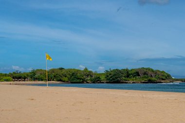 Sarı bayrak sahilde, bir su kütlesinin yanında. Bayrak bir direğin üzerinde ve ön planda. Kumsal boş ve gökyüzü temiz.