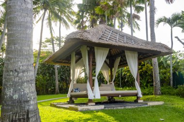 Beyaz perdeleri ve sazdan çatısı olan küçük bir kulübe. Kulübe ağaçlar ve otlarla çevrili.