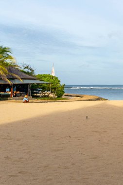 Bali, Endonezya - 19 Aralık 2024: küçük bir kulübesi ve kumsalda oturan birkaç kişi. Kulübe suya yakın ve sahil boş.