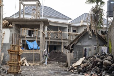 Bali, Endonezya - 25 Aralık 2024: inşaat sahasında sert şapkalı ve mavi tenteli bir adam var. Adam sitenin etrafında dolaşıyor, muhtemelen ilerlemeyi inceliyor ya da her şeyin güvende olduğundan emin oluyor.