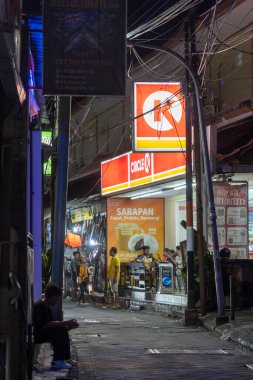 Bali, Endonezya - 25 Aralık 2024: Adam Circle K süpermarketinin önündeki kaldırımda oturuyor. Sokak karanlık ve adam sarı bir gömlek giyiyor.