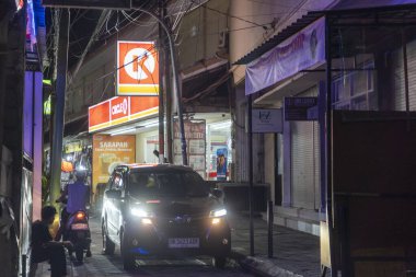 Bali, Endonezya - 25 Aralık 2024: Adam Circle K süpermarketinin önündeki kaldırımda oturuyor. Sokak karanlık ve adam sarı bir gömlek giyiyor.