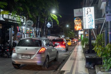 Bali, Endonezya - 25 Aralık 2024: Bir sürü arabası ve bir kadın reklam panosu olan işlek cadde. Sokaklar geceleri aydınlık.