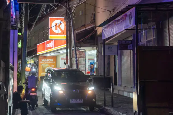 Bali, Endonezya - 25 Aralık 2024: Adam Circle K süpermarketinin önündeki kaldırımda oturuyor. Sokak karanlık ve adam sarı bir gömlek giyiyor.