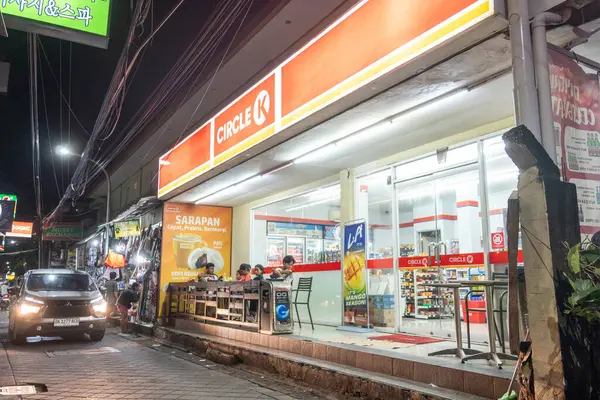 Bali, Endonezya - 25 Aralık 2024: Adam Circle K süpermarketinin önündeki kaldırımda oturuyor. Sokak karanlık ve adam sarı bir gömlek giyiyor.