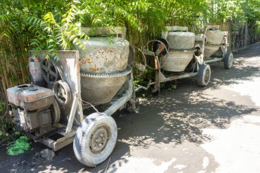 Paslı tekerleği olan çimento karıştırıcısı bahçede duruyor. Tekerlek kayıp ve makine bakımsız.